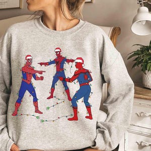 Może przedstawiać: Jasnoszara bluza z nadrukiem przedstawiającym trzech Spider-Manów w czapkach Świętego Mikołaja i zaplątanych w lampki choinkowe. Postacie Spider-Mana są w swoich klasycznych czerwono-niebieskich kostiumach.