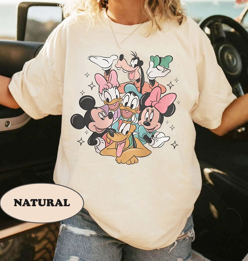 May include: A white t-shirt with a graphic print of Mickey Mouse, Minnie Mouse, Donald Duck, Goofy, and Pluto. The characters are in a playful pose with stars surrounding them. The text 'NATURAL' is printed on the bottom left corner of the shirt.