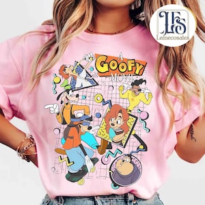 May include: Pink t-shirt featuring a colorful graphic from "A Goofy Movie." The design includes cartoon characters Goofy, Max, and others, with the movie title in bold letters. The background has a grid pattern with abstract shapes.
