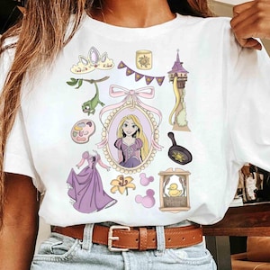 以下が含まれることがあります： 白いTシャツに漫画のイラストが描かれています。デザインには、王女の肖像画、塔、フライパン、ドレス、その他の関連要素が、紫、ピンク、黄色の色合いで描かれています。