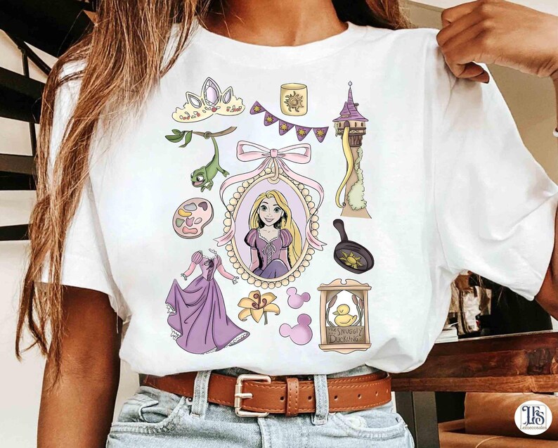 以下が含まれることがあります： 白いTシャツに漫画のイラストが描かれています。デザインには、王女の肖像画、塔、フライパン、ドレス、その他の関連要素が、紫、ピンク、黄色の色合いで描かれています。