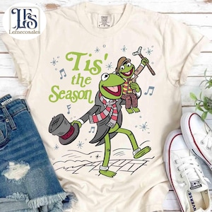 Puede incluir: Camiseta color crema con la Rana Gustavo en una escena invernal con el texto "Tis' the Season". Gustavo lleva un sombrero de copa, bufanda y abrigo, con un amigo. Copos de nieve y notas musicales decoran el diseño.