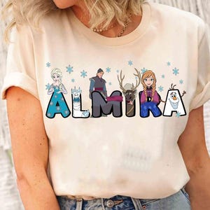 Puede incluir: Una camiseta blanca con un gr&aacute;fico de los personajes de la pel&iacute;cula Frozen. El gr&aacute;fico incluye a Elsa, Anna, Olaf, Kristoff y Sven. Los personajes est&aacute;n dispuestos para deletrear el nombre "ALMIRA".