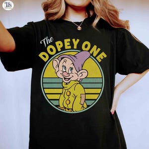 May include: Black t-shirt featuring a cartoon image of Dopey from Snow White. The image is in a circular design with the text "The DOPEY ONE". Dopey is wearing a purple hat and yellow outfit.