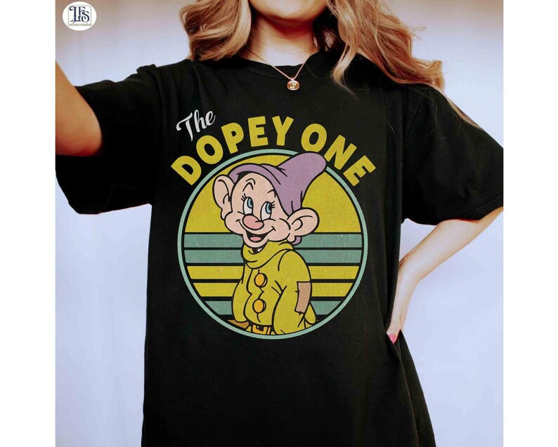 May include: Black t-shirt featuring a cartoon image of Dopey from Snow White. The image is in a circular design with the text "The DOPEY ONE". Dopey is wearing a purple hat and yellow outfit.