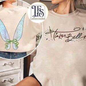 May include: Beige sweatshirt featuring a graphic of Tinker Bell's wings on the back and her name in cursive on the front. The wings are light blue and green with a green bow. The text "Tinker Bell" is in dark brown.