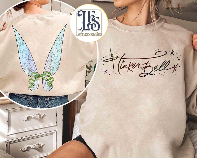 May include: Beige sweatshirt featuring a graphic of Tinker Bell's wings on the back and her name in cursive on the front. The wings are light blue and green with a green bow. The text "Tinker Bell" is in dark brown.