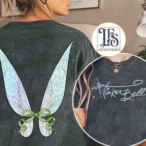 May include: Dark gray t-shirt featuring a large, colorful fairy wing design on the back. The wings are light blue, green, and white with a green bow. The front of the shirt has the words "Tinker Bell" in a script font.