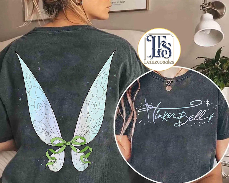 May include: Dark gray t-shirt featuring a large, colorful fairy wing design on the back. The wings are light blue, green, and white with a green bow. The front of the shirt has the words "Tinker Bell" in a script font.