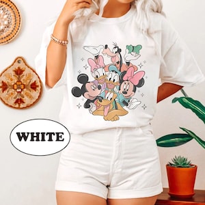 May include: White t-shirt with a graphic print of Mickey Mouse, Minnie Mouse, Donald Duck, Goofy, and Pluto. The characters are in a playful pose, with Mickey Mouse holding Pluto's tail. The shirt has a crew neck and short sleeves.