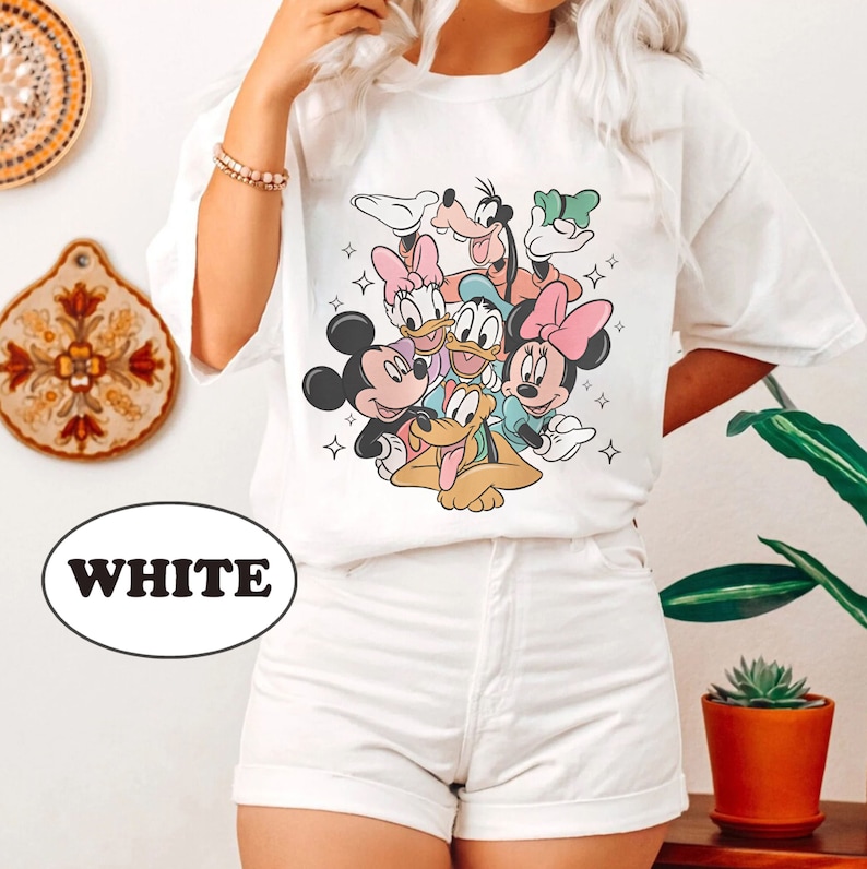 May include: White t-shirt with a graphic print of Mickey Mouse, Minnie Mouse, Donald Duck, Goofy, and Pluto. The characters are in a playful pose, with Mickey Mouse holding Pluto's tail. The shirt has a crew neck and short sleeves.