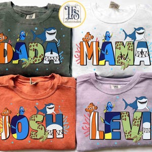 May include: Four crewneck sweatshirts in green, white, orange, and lavender. Each shirt features a colorful design with the words "DADA", "MAMA", "JOSH", and "LEVI", along with cartoon characters of a clownfish, a blue tang, and a shark.