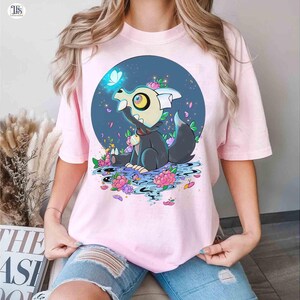 K&ouml;nnte beinhalten: Hellrosa T-Shirt mit einem Cartoon-Charakter mit gro&szlig;em Auge, der in einer Blumenszene sitzt. Der Charakter ist von Blumen und einem blauen Kreis-Hintergrund umgeben. Das T-Shirt besteht aus einem weichen Material.
