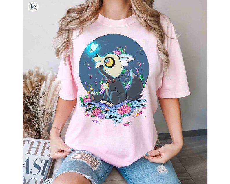 K&ouml;nnte beinhalten: Hellrosa T-Shirt mit einem Cartoon-Charakter mit gro&szlig;em Auge, der in einer Blumenszene sitzt. Der Charakter ist von Blumen und einem blauen Kreis-Hintergrund umgeben. Das T-Shirt besteht aus einem weichen Material.