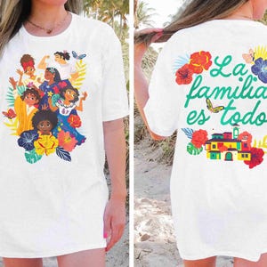 May include: White t-shirt featuring a colorful illustration of animated characters surrounded by flowers and butterflies. The back of the shirt has the text "La familia es todo" with a butterfly and floral accents. A casual, comfortable style.