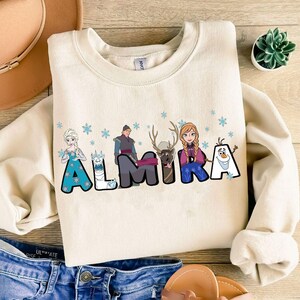 Puede incluir: Una sudadera de color crema con un gr&aacute;fico de personajes de la pel&iacute;cula Frozen. Los personajes est&aacute;n dispuestos para deletrear el nombre "ALMIRA".