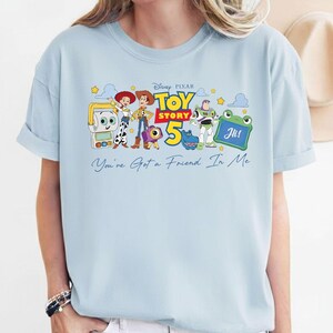 May include: Light blue t-shirt featuring a graphic of Toy Story 5 characters. The design includes Woody, Buzz Lightyear, and other characters, with the text "Toy Story 5" and "You've Got a Friend In Me".