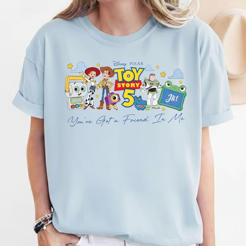 May include: Light blue t-shirt featuring a graphic of Toy Story 5 characters. The design includes Woody, Buzz Lightyear, and other characters, with the text "Toy Story 5" and "You've Got a Friend In Me".