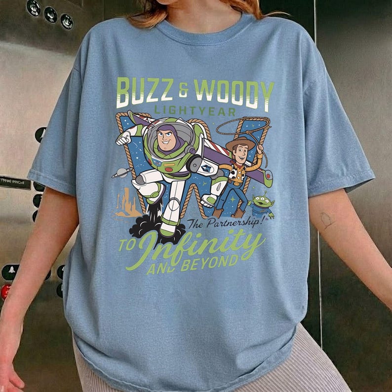 May include: Light blue t-shirt featuring a graphic of Buzz Lightyear and Woody from Toy Story. The design includes the text "Buzz & Woody Lightyear" and "To Infinity and Beyond". The shirt is a relaxed fit.
