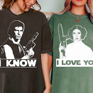 May include: Two t-shirts, one dark grey and one green, with black and white images of characters holding guns. The dark grey shirt has the text "I KNOW", and the green shirt says "I LOVE YOU".
