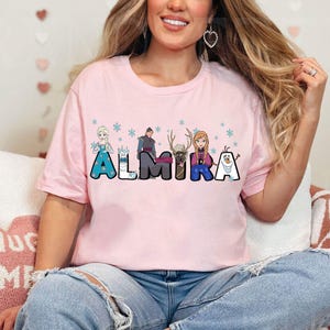 Puede incluir: Camiseta rosa con un gr&aacute;fico de personajes de la pel&iacute;cula Frozen. Los personajes est&aacute;n dispuestos para deletrear el nombre "Almira".