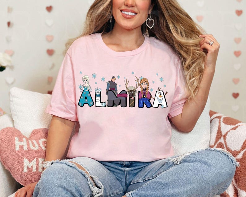 Puede incluir: Camiseta rosa con un gr&aacute;fico de personajes de la pel&iacute;cula Frozen. Los personajes est&aacute;n dispuestos para deletrear el nombre "Almira".