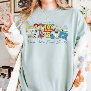 May include: A light blue sweatshirt featuring a colorful graphic of characters from the Disney Pixar movie Toy Story 5. The graphic includes Woody, Buzz Lightyear, and other characters, along with the text "Toy Story 5" and "You've Got a Friend In Me".