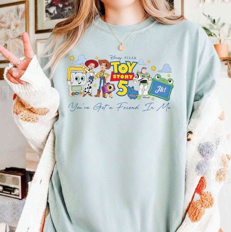 May include: A light blue sweatshirt featuring a colorful graphic of characters from the Disney Pixar movie Toy Story 5. The graphic includes Woody, Buzz Lightyear, and other characters, along with the text "Toy Story 5" and "You've Got a Friend In Me".