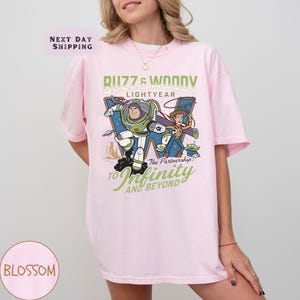 May include: A light pink t-shirt featuring a graphic of Buzz Lightyear and Woody from Toy Story. The design includes the text "Buzz & Woody Lightyear" and "To Infinity and Beyond". The shirt is a comfortable, casual style.