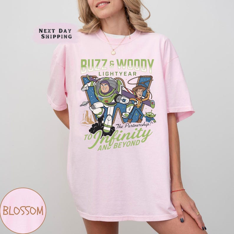 May include: A light pink t-shirt featuring a graphic of Buzz Lightyear and Woody from Toy Story. The design includes the text "Buzz & Woody Lightyear" and "To Infinity and Beyond". The shirt is a comfortable, casual style.