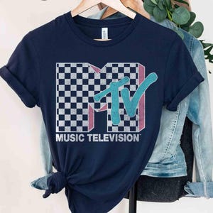 May include: Navy blue t-shirt featuring the MTV logo with a checkered pattern, pink accents, and a teal "TV" design. The words "MUSIC TELEVISION" are printed below the logo in white. The shirt is hanging on a wooden hanger.