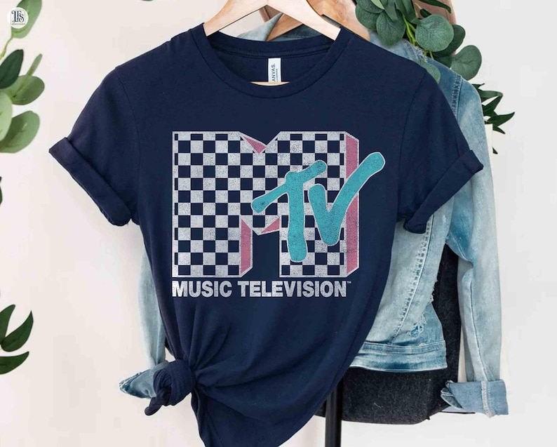 May include: Navy blue t-shirt featuring the MTV logo with a checkered pattern, pink accents, and a teal "TV" design. The words "MUSIC TELEVISION" are printed below the logo in white. The shirt is hanging on a wooden hanger.