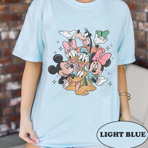 May include: Light blue t-shirt featuring a graphic print of Mickey Mouse, Minnie Mouse, Donald Duck, Goofy, and Pluto. The characters are arranged in a circle with their arms around each other. The shirt has a crew neck and short sleeves.
