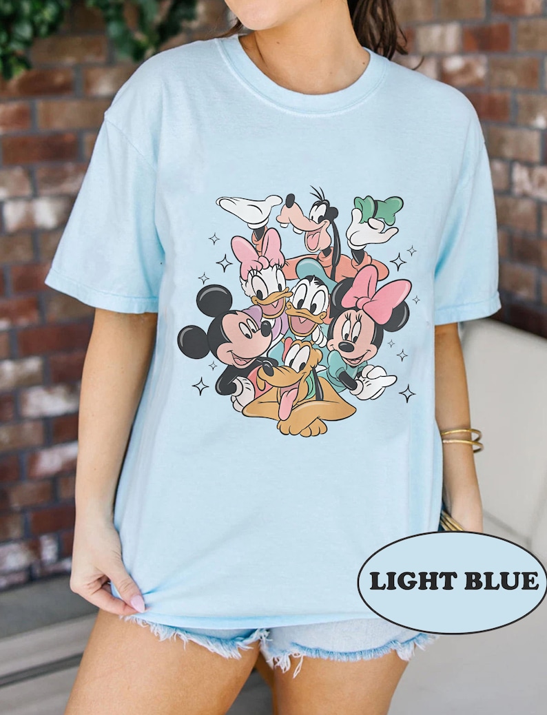 May include: Light blue t-shirt featuring a graphic print of Mickey Mouse, Minnie Mouse, Donald Duck, Goofy, and Pluto. The characters are arranged in a circle with their arms around each other. The shirt has a crew neck and short sleeves.