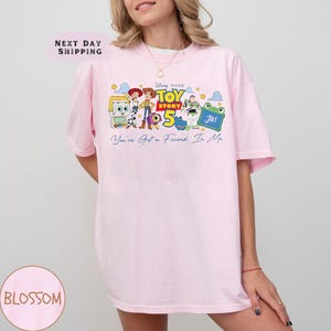 May include: A light pink t-shirt featuring a colorful graphic of characters from the Disney Pixar movie Toy Story 5. The design includes the text "Toy Story 5" and "You've Got a Friend In Me". The shirt also has the text "Next Day Shipping".