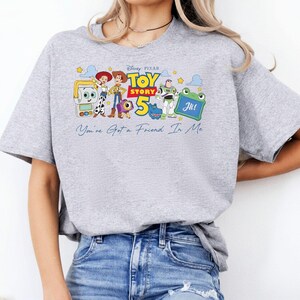 May include: A light gray t-shirt featuring a colorful graphic of characters from the Disney Pixar movie Toy Story 5. The graphic includes the text "Toy Story 5" and "You've Got a Friend In Me".