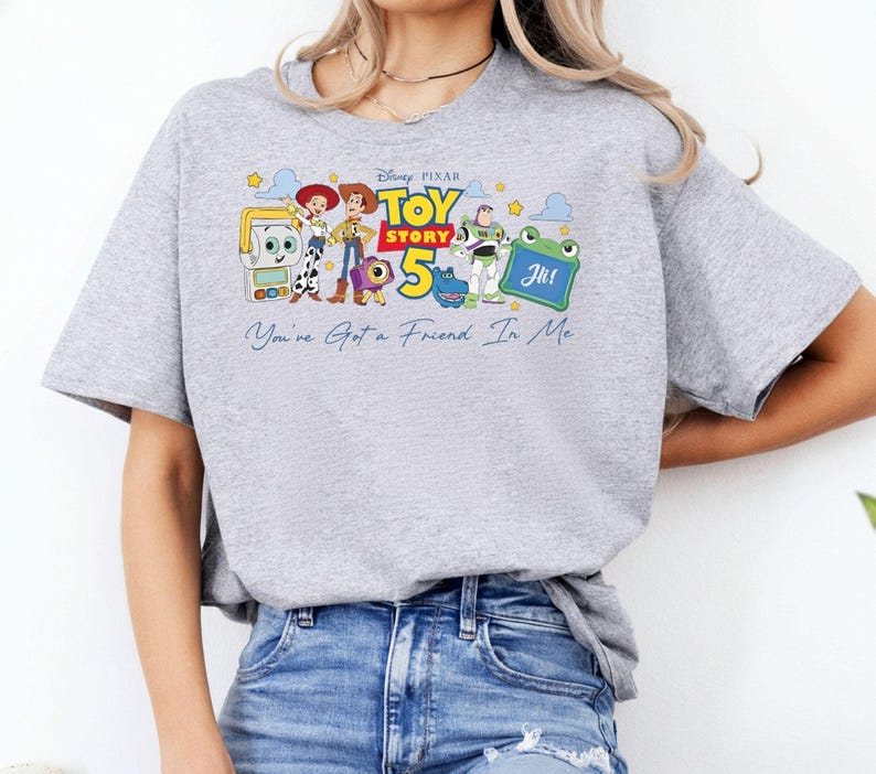 May include: A light gray t-shirt featuring a colorful graphic of characters from the Disney Pixar movie Toy Story 5. The graphic includes the text "Toy Story 5" and "You've Got a Friend In Me".