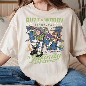 May include: Cream-colored t-shirt featuring a graphic of Buzz Lightyear and Woody from Toy Story. The design includes the text "Buzz & Woody Lightyear" and "To Infinity and Beyond." The characters are depicted in a dynamic pose with a desert landscape backdrop.