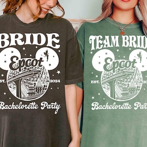 Può includere: Due magliette, una grigio scuro e una verde, con testo e grafica bianchi. La maglietta grigio scuro dice "BRIDE Epcot Bachelorette Party EST. 2024". La maglietta verde dice "TEAM BRIDE Epcot Bachelorette Party EST. 2024". Entrambe le magliette hanno un design a testa di Topolino.