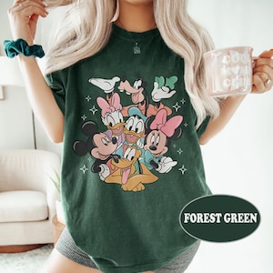 May include: A forest green t-shirt with a graphic print of Mickey Mouse, Minnie Mouse, Donald Duck, Goofy, and Pluto. The characters are arranged in a circle with their heads facing the center. The shirt has a relaxed fit and a crew neck.