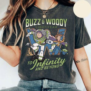 May include: Dark gray t-shirt featuring Buzz Lightyear and Woody from Toy Story. The graphic includes the text "Buzz & Woody Lightyear" and "To Infinity and Beyond". The design shows the characters in action with a space and desert background.