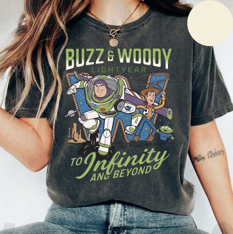 May include: Dark gray t-shirt featuring Buzz Lightyear and Woody from Toy Story. The graphic includes the text "Buzz & Woody Lightyear" and "To Infinity and Beyond". The design shows the characters in action with a space and desert background.