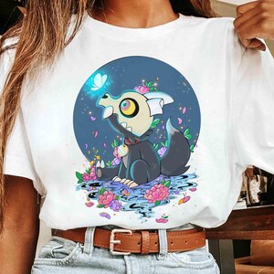 K&ouml;nnte beinhalten: Wei&szlig;es T-Shirt mit einer Cartoon-Illustration einer schwarz-grauen Kreatur mit Krone, umgeben von Blumen und einem Schmetterling. Das Design befindet sich vor einem blauen kreisf&ouml;rmigen Hintergrund.