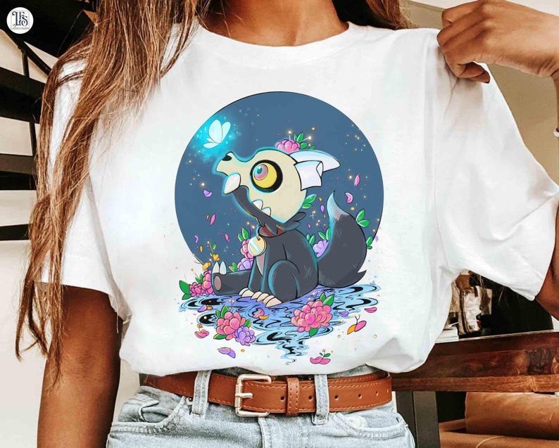 K&ouml;nnte beinhalten: Wei&szlig;es T-Shirt mit einer Cartoon-Illustration einer schwarz-grauen Kreatur mit Krone, umgeben von Blumen und einem Schmetterling. Das Design befindet sich vor einem blauen kreisf&ouml;rmigen Hintergrund.