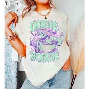 以下が含まれることがあります： クリーム色のTシャツ。緑と紫で「Oogie Boogie」の文字とキャラクターのグラフィックが描かれています。キャラクターはサイコロを持っています。Tシャツは半袖で、クルーネックです。