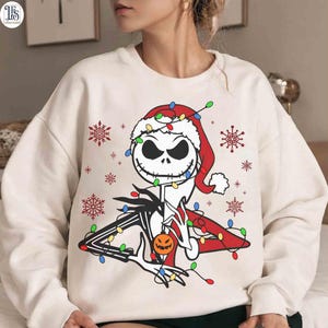Puede incluir: Sudadera crema con un dise&ntilde;o de Jack Skellington con gorro de Pap&aacute; Noel, luces navide&ntilde;as y copos de nieve. El dise&ntilde;o incluye una calabaza, fusionando Halloween y Navidad en un estilo festivo.
