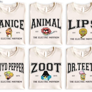 Könnte beinhalten: Sechs cremefarbene T-Shirts mit verschiedenen Muppets-Charakteren. Jedes Shirt hat den Namen eines Charakters in schwarzer Schrift im College-Stil, zusammen mit dem Text "EST 1975" und "THE ELECTRIC MAYHEM". Zu den Charakteren gehören Janice, Animal, Lips, Floyd Pepper, Zoot und Dr. Teeth.