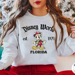 May include: A light gray sweatshirt with the text "Disney World" above an image of Mickey Mouse wearing a Santa hat and Christmas lights. The text "est. 1971" and "FLORIDA" are also printed on the sweatshirt.