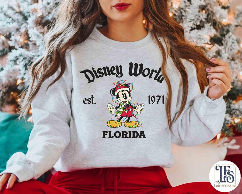 May include: A light gray sweatshirt with the text "Disney World" above an image of Mickey Mouse wearing a Santa hat and Christmas lights. The text "est. 1971" and "FLORIDA" are also printed on the sweatshirt.