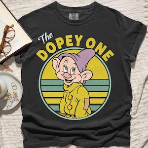 May include: Black t-shirt featuring a cartoon image of Dopey from Snow White. The graphic shows Dopey in a yellow outfit and purple hat, set against a yellow and blue striped circle. The text "The Dopey One" is above the character.